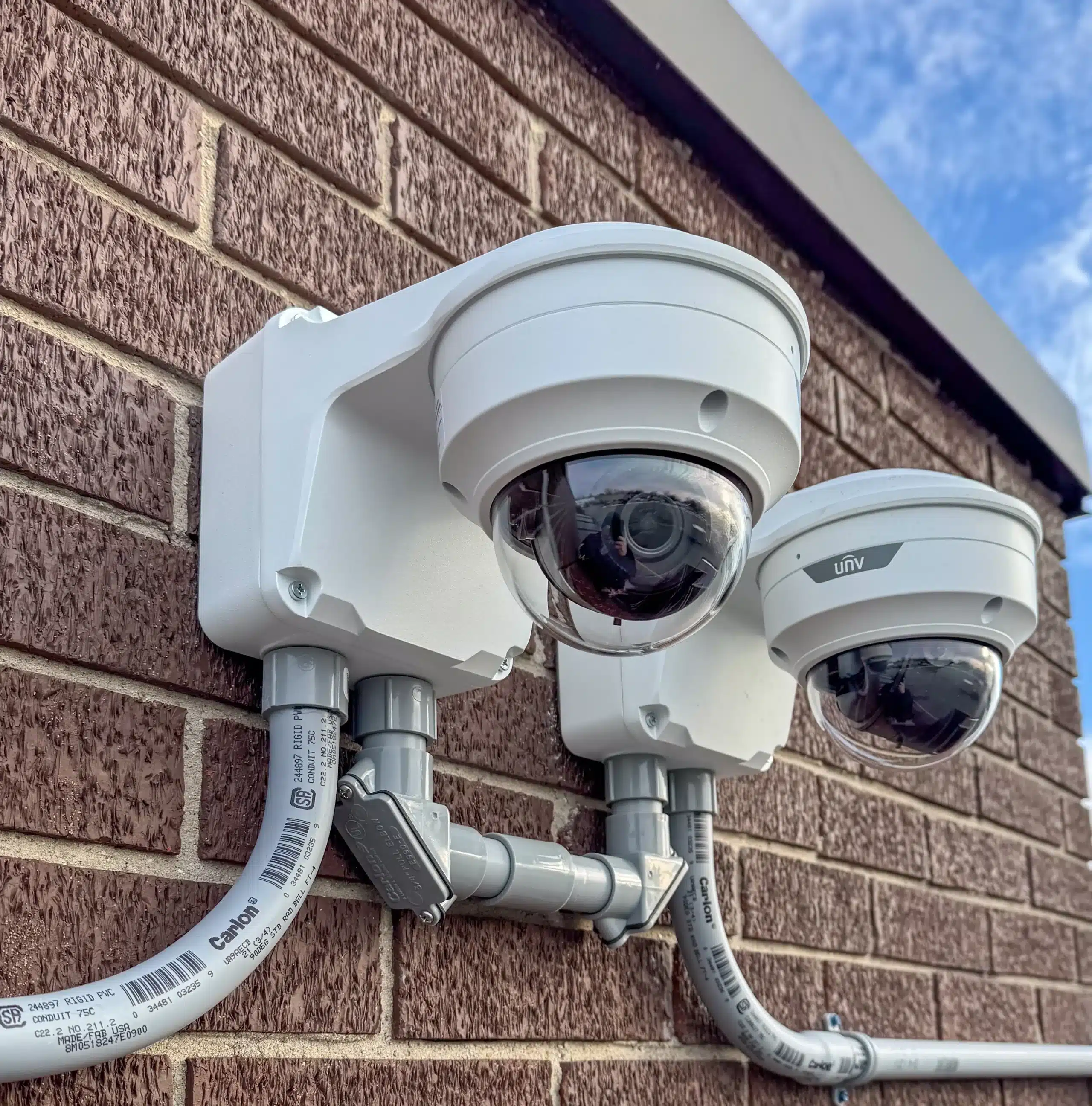Two dome security cameras mounted on a brick wall, connected by exposed electrical conduits, under a partly cloudy sky.