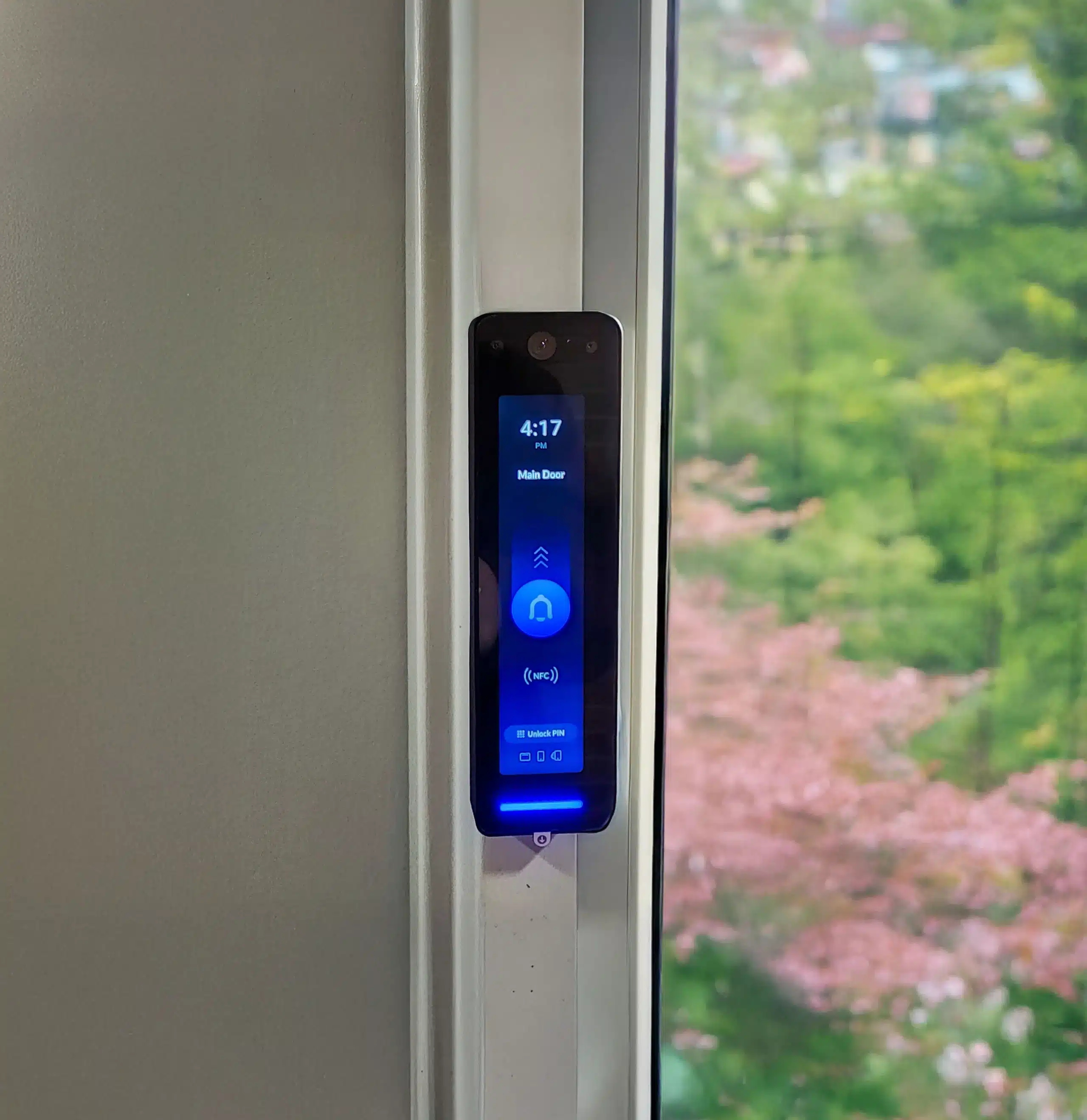 A digital door lock with a touchscreen display is mounted on a white door frame, showing the time and lock status. A blurred garden is visible outside the window.